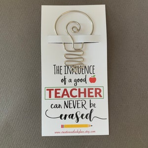 May include: A white card with a silver wire lightbulb design and the text "The influence of a good teacher can never be erased."  The card also includes a red apple and the website address "www.creativeoutlookglass.etsy.com".