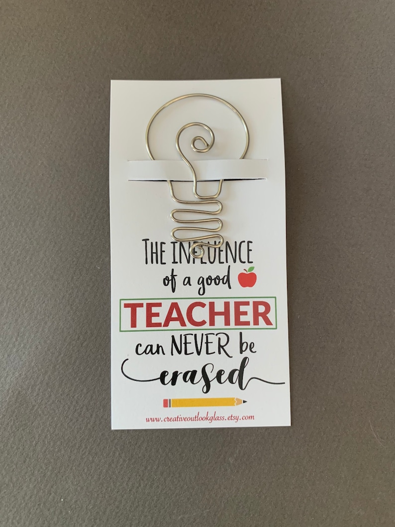 May include: A white card with a silver wire lightbulb design and the text "The influence of a good teacher can never be erased."  The card also includes a red apple and the website address "www.creativeoutlookglass.etsy.com".