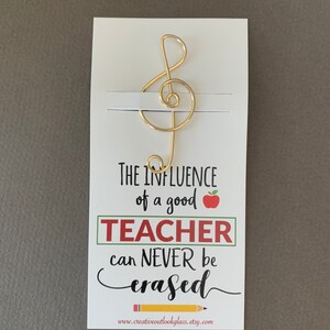 May include: A white card with a gold treble clef bookmark attached. The card reads "The influence of a good teacher can never be erased." with a red apple and a yellow pencil graphic. The website www.creativeoutlookglass.etsy.com is printed at the bottom.