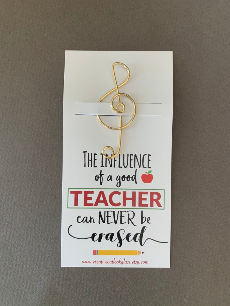 May include: A white card with a gold treble clef bookmark attached. The card reads "The influence of a good teacher can never be erased." with a red apple and a yellow pencil graphic. The website www.creativeoutlookglass.etsy.com is printed at the bottom.