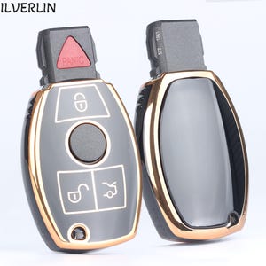 May include: Two black car key fobs with gold trim. One fob has a panic button and four buttons, including a lock, unlock, and trunk button. The other fob has a smooth, black surface.