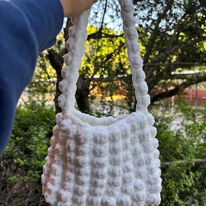May include: White crocheted handbag with a fluffy texture and a long strap. The bag is made of a soft, white yarn and has a simple, elegant design.