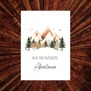 Könnte beinhalten: Ein weißer rechteckiger Druck mit einer Aquarellillustration von Bergen, Bäumen und Vögeln. Der Text "AUF INS NÄCHSTE Abenteuer" ist in schwarzer Schrift geschrieben. Der Druck wird auf einer Holzoberfläche angezeigt.