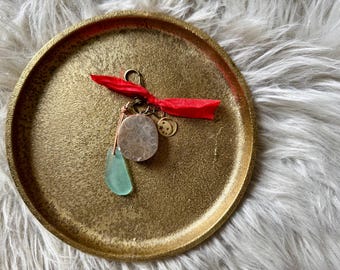 May include: A gold-colored metal tray with a textured surface holds a collection of charms. A red ribbon is tied to a keychain with a coral-like stone, a green teardrop-shaped charm, and a gold moon charm.