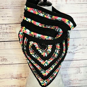 May include: A black crochet cowl scarf with colorful floral accents. The scarf is displayed on a mannequin and features a triangular shape with a heart-shaped design at the bottom. The colorful accents include pink, yellow, and blue.