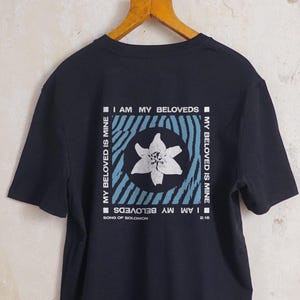 Pode incluir: Camiseta preta com um design floral branco e o texto "I AM MY BELOVEDS" em uma moldura quadrada. A camisa também apresenta o texto "MY BELOVED IS MINE" nas laterais. A camisa está pendurada em um cabide de madeira.