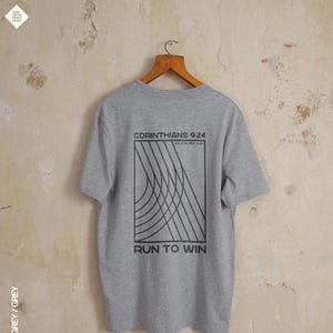May include: Grey t-shirt with a black graphic on the back. The graphic features the text "CORINTHIANS 9:24" above curved lines and the words "RUN TO WIN" below. The shirt is displayed on a wooden hanger.