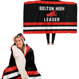 Pode incluir: Uma manta com capuz preta e vermelha com listras brancas. A manta tem o texto "Bolton High Cheer Leader" com um gráfico de megafone vermelho.