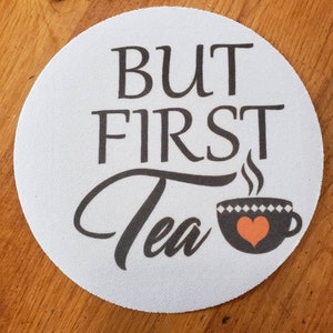 May include: White round coaster with the text "BUT FIRST Tea" in black lettering. A black and white teacup with a red heart is shown next to the text.