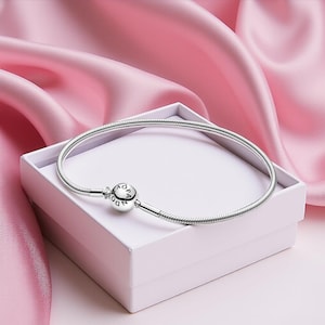 May include: A silver bracelet with a round clasp, resting on a white box. The bracelet has a snake chain design and is displayed against a backdrop of pink fabric. The clasp features the word "PANDORA".