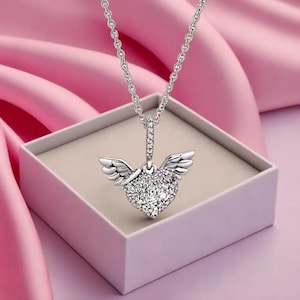 Op de afbeelding: Een zilveren ketting met een hartvormige hanger versierd met sprankelende kristallen en kleine vleugels. De ketting wordt gepresenteerd in een witte doos tegen een roze achtergrond, wat het delicate ontwerp van de sieraden benadrukt.