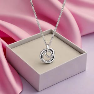 May include: A silver necklace with a pendant of two intertwined circles, one set with small, clear gemstones. The necklace is presented in a light beige box, set against a backdrop of pink fabric.