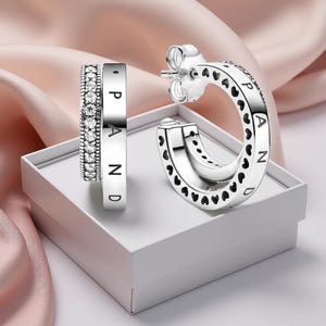 Könnte beinhalten: Silberne Creolen mit Herz-Ausschnitten und dem eingravierten Wort "PAND". Ein Ohrring hat eine Reihe kleiner, klarer Steine. Die Ohrringe werden auf einer weißen Schachtel vor rosa Hintergrund präsentiert.