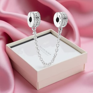 Può includere: Una catena di sicurezza Pandora argentata con due charm cilindrici, ciascuno ornato di pietre scintillanti e il nome del marchio. Gli charm sono collegati da una delicata catena, presentati su una scatola bianca con il logo Pandora, su sfondo di raso rosa.