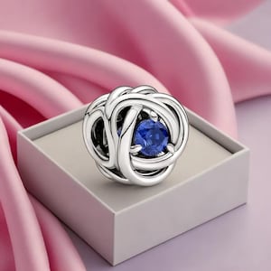 May include: A silver charm with an intricate, interwoven design, featuring a vibrant blue gemstone at its center. The charm is presented on a light gray box, set against a backdrop of soft pink fabric, creating a visually appealing contrast.
