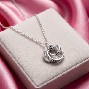 May include: A silver necklace featuring two intertwined circles. One circle is set with small, clear stones, the other with a twisted rope pattern. The necklace is displayed on a light-coloured, square surface, against a pink satin backdrop.