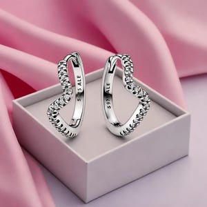 May include: A pair of silver heart-shaped hoop earrings. Each earring is encrusted with small, clear gemstones along the outer curve. The earrings are displayed in a white box on a pink surface. The text "S925 ALE" is visible on each earring.