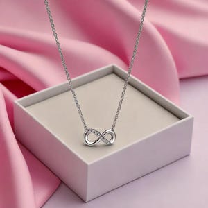 Könnte beinhalten: Eine silberfarbene Halskette mit einem Unendlichkeitszeichen-Anhänger. Der Anhänger ist mit kleinen, klaren Edelsteinen besetzt. Die Halskette wird auf einer weißen Schachtel vor rosa Hintergrund präsentiert.