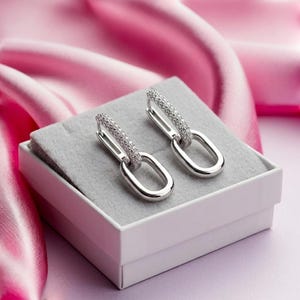 May include: A pair of silver-toned hoop earrings featuring a dangling oval link design. The upper portion of each earring is encrusted with small, clear, sparkling stones, while the lower oval link remains a smooth, polished silver-toned metal.