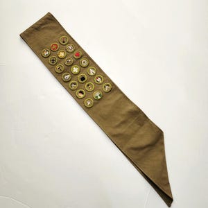 May include: A khaki-colored merit badge sash with multiple embroidered patches. The patches feature various designs, including a red heart, a white cross, and a tent. The sash is made of a durable fabric and is designed to be worn across the chest.