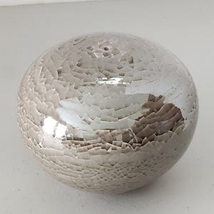 May include: A decorative, spherical object with a cracked, mosaic-like surface. The color is a mix of silver and light gray, with a glossy finish. The sphere has a smooth, rounded shape, and the surface appears to be made of small, irregular pieces.