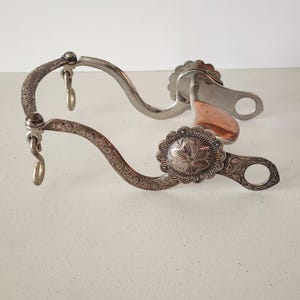 May include: A silver-toned metal horse bit featuring intricate floral scrollwork engravings on the curved shanks. The bit includes a copper-coloured mouthpiece and two circular conchos decorated with a central star pattern and scalloped edges.