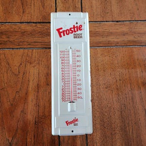 May include: A vintage white metal thermometer for Frostie Root Beer. The thermometer has a red and white temperature scale, with the Frostie logo at the top and bottom. The thermometer measures in Fahrenheit and Celsius.