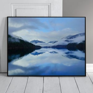 May include: A framed landscape print featuring a serene lake scene. The image captures a mountain range shrouded in mist, reflected in the calm water. The sky is a mix of blue and cloudy white, creating a tranquil and natural aesthetic.