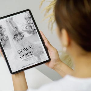 Op de afbeelding: Een tablet met een zwart-wit afbeelding van een trouwjurk met de tekst "GOWN GUIDE" in een schreefletter. De tablet wordt door een persoon vastgehouden, met de focus op het scherm. De achtergrond is wazig.