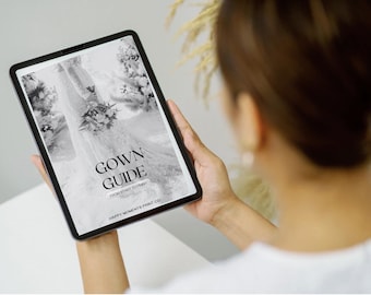 May include: A tablet displaying a black and white image of a wedding gown with the text "GOWN GUIDE" in a serif font. The tablet is held by a person, with the focus on the screen. The background is blurred.
