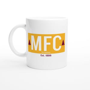Puede incluir: Taza de cerámica blanca con un rectángulo amarillo con las letras "MFC" en blanco. El texto "The Steelmen" y "Est. 1886" están debajo. Dos pinos granates flanquean las letras.