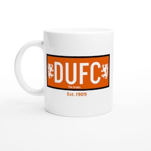 May include: White ceramic mug featuring an orange rectangle with the text "DUFC" in white, flanked by lion emblems. Below, the text "The Arabs" and "Est. 1909" are printed in black. The mug has a curved handle.
