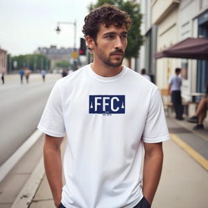 以下が含まれることがあります： 前面に紺色の長方形のグラフィックが入った白いTシャツ。グラフィックには白い文字で「FFC」と、その下に「Est. 1876」の文字が描かれています。Tシャツは半袖です。