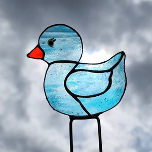 Puede incluir: Una figura de pato de vidriera con cuerpo azul claro, contornos negros y pico naranja. El ave tiene un ojo pequeño y oscuro y está montada sobre una estaca de metal negro para la decoración del jardín.
