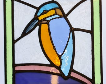 Könnte beinhalten: Ein Buntglasfenster mit einem blauen und orangefarbenen Eisvogel, der auf einem Ast sitzt. Der Vogel ist schwarz umrandet und der Hintergrund ist hellblau.