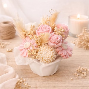 May include: A floral arrangement featuring pink rose-shaped soaps, light pink daisies, and a cream-colored chrysanthemum. The arrangement is in a white ceramic container, with dried grasses and a lit candle in the background.