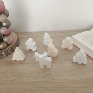 May include: A collection of white and off-white Christmas-themed decorations. The set includes a gingerbread man, snowman, Christmas tree, reindeer, and house-shaped ornaments. The decorations are arranged on a wooden surface, with a book and Christmas ornaments in the background.