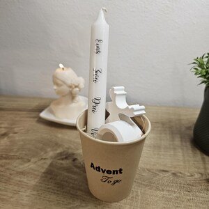 Könnte beinhalten: Eine weiße Adventskerze mit schwarzer Schrift in einem kleinen braunen Pappbecher. Auf dem Becher steht "Advent To go". Im Hintergrund brennt eine weiße Kerze in Form einer Frauenbüste.