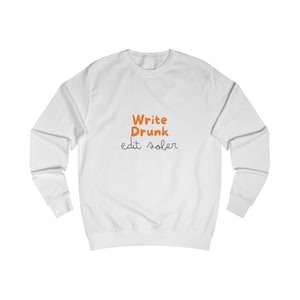 Puede incluir: Sudadera blanca con mangas largas. La parte delantera de la sudadera presenta el texto "Write Drunk edit sober" en naranja y negro. La sudadera está hecha de un material suave y cómodo.