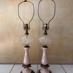 May include: A pair of table lamps featuring pale pink ceramic bases with ornate bronze-toned metal accents at the bottom. Each lamp has a clear, textured glass globe section above the ceramic base, topped with a brass-toned metal harp fixture.