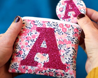 May include: A pink floral fabric pouch with a large pink glitter letter A on the front. A smaller pink glitter letter A is attached to the pouch.