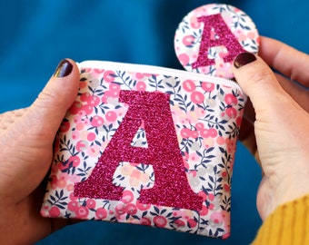 May include: A pink and white floral fabric pouch with a zipper closure. The pouch has a large pink glitter letter "A" on the front.