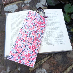 May include: A white fabric glasses case with a pink and blue floral pattern. The case is lying on top of a book.