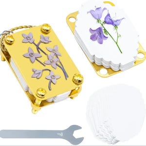 May include: A gold-colored metal book-shaped box with a floral design, a stack of white scalloped-edge tags, and a silver wrench. The box has a braided handle and is adorned with gold screws. A separate gold holder contains tags with purple flower illustrations.