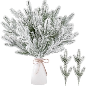 May include: A white ceramic vase holds a bundle of faux evergreen branches dusted with white snow. A twine bow adorns the vase. Two additional snow-covered branches are displayed to the right, suggesting a winter holiday theme.