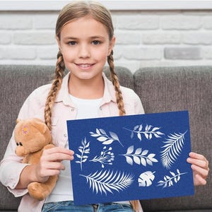 Może przedstawiać: Młoda dziewczyna trzyma niebieską kartkę z białymi wzorami liści. Karta zawiera r&oacute;żne botaniczne ilustracje liści i liści. Trzyma r&oacute;wnież misia. Tło obejmuje szarą sofę i białą ceglaną ścianę.