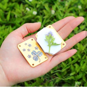 以下が含まれることがあります： 表面に四つ葉のクローバーと小さな花が押し付けられた、金色の金属製の正方形が2つ。正方形は角に穴が開いています。正方形は緑色の背景に対して手で持たれています。