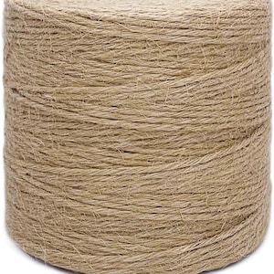 May include: A large, cylindrical spool of natural tan jute twine. The thick, fibrous cord is tightly wound in a continuous spiral pattern, creating a textured surface with visible loose fibers and a consistent light brown, earthy color throughout.