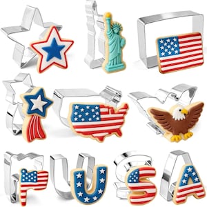 May include: A collection of cookie cutters and decorated cookies in patriotic themes. Shapes include a star, the Statue of Liberty, the American flag, the USA map, an eagle, and the letters U, S, and A. The cookies are decorated with red, white, and blue icing.