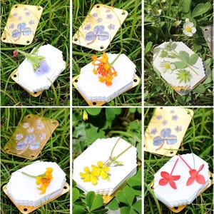 以下が含まれることがあります： 白い紙に様々な花や葉が押し花された、6つのしおり。しおりは緑の芝生の上に置かれ、金色の金属フレームに取り付けられています。